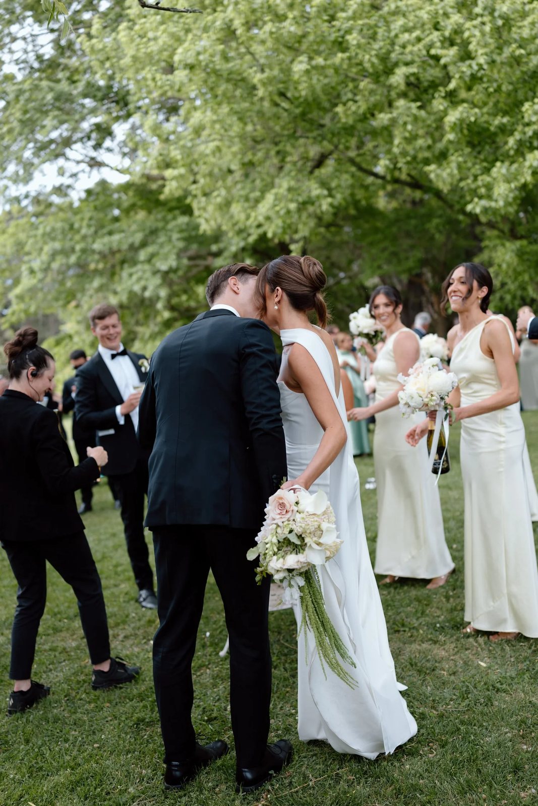 A couple kissing on a lawn as the bridesmaids look on, bouquets in hand
