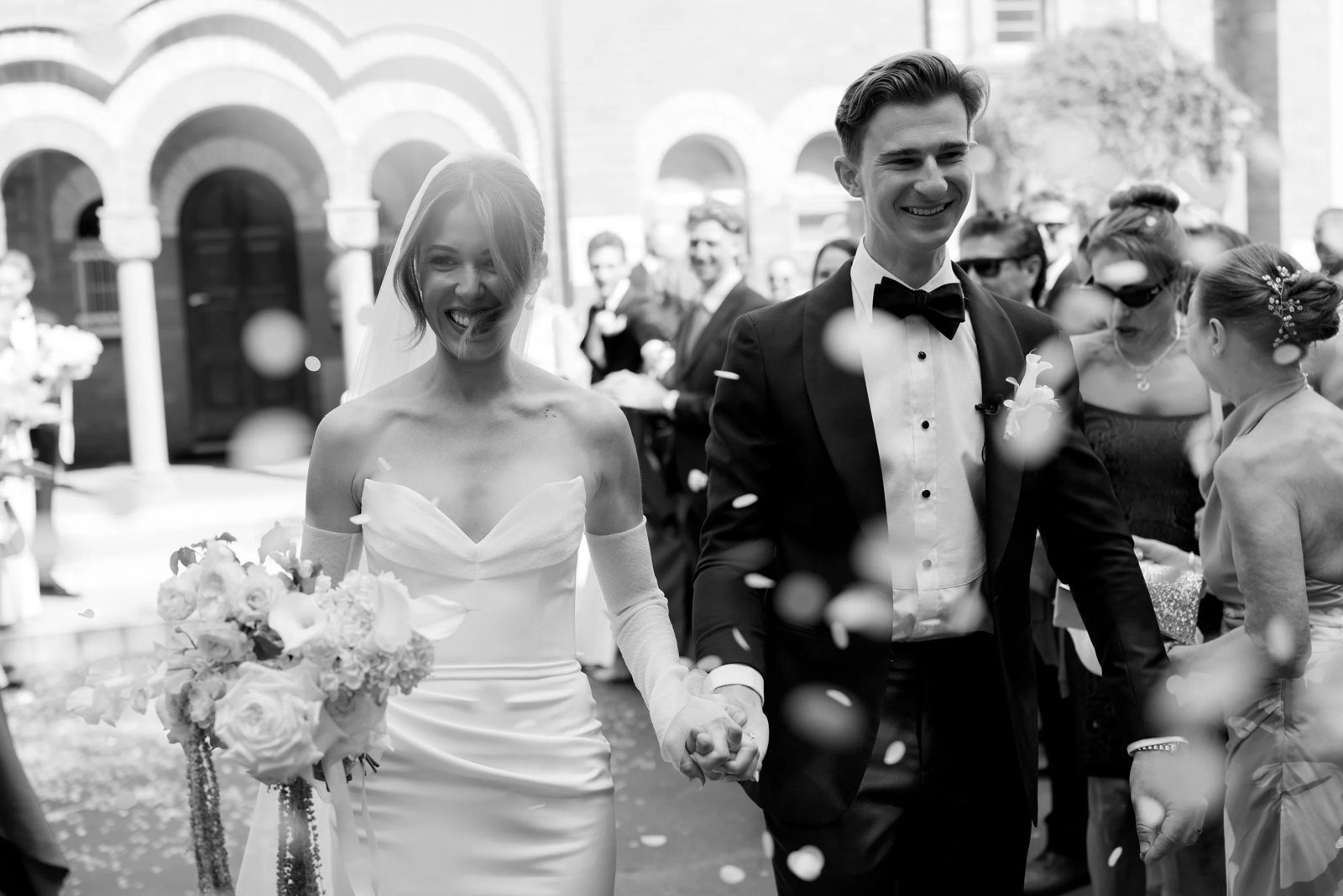 Black and white: a couple laughing hand in hand through falling confetti outside a church