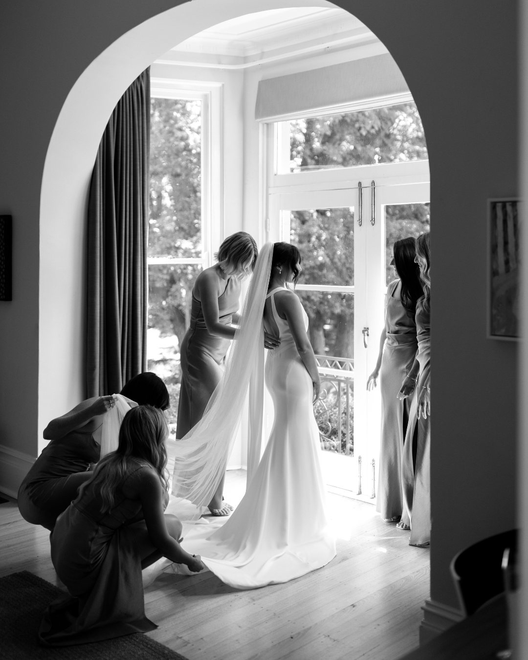 Black and white: bridesmaids fastening a bride&rsquo;s gown in an arched doorway