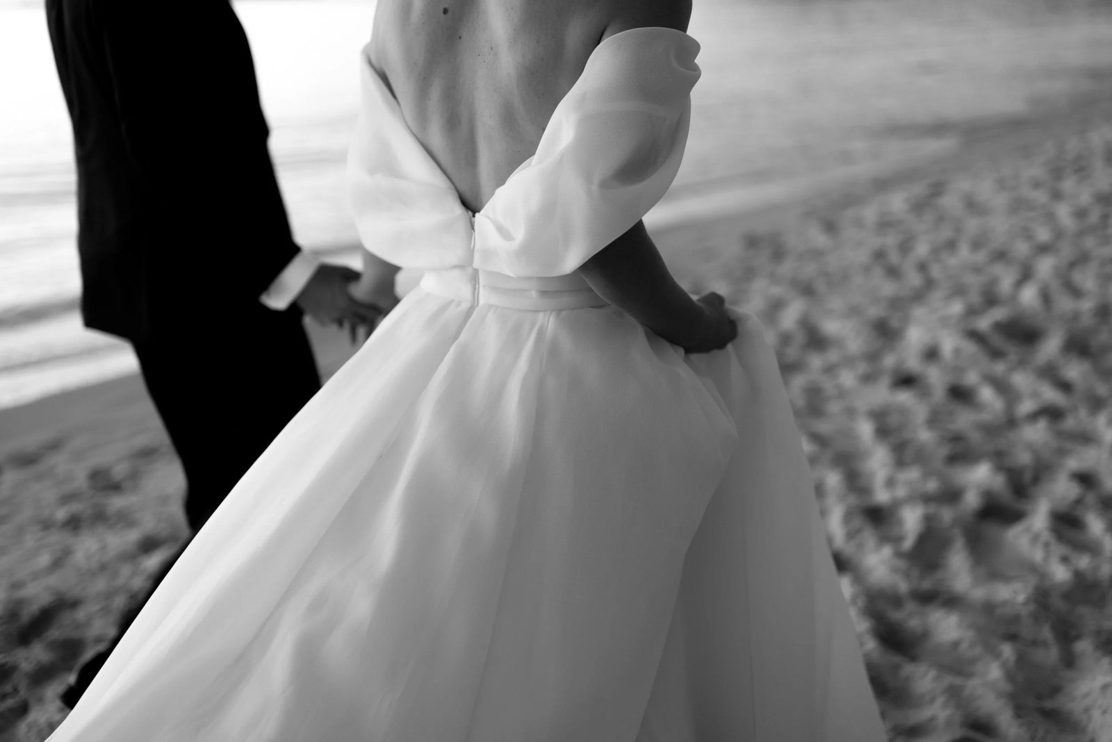 Black and white: the back of a wedding gown on the sand, the groom&rsquo;s hand at her waist