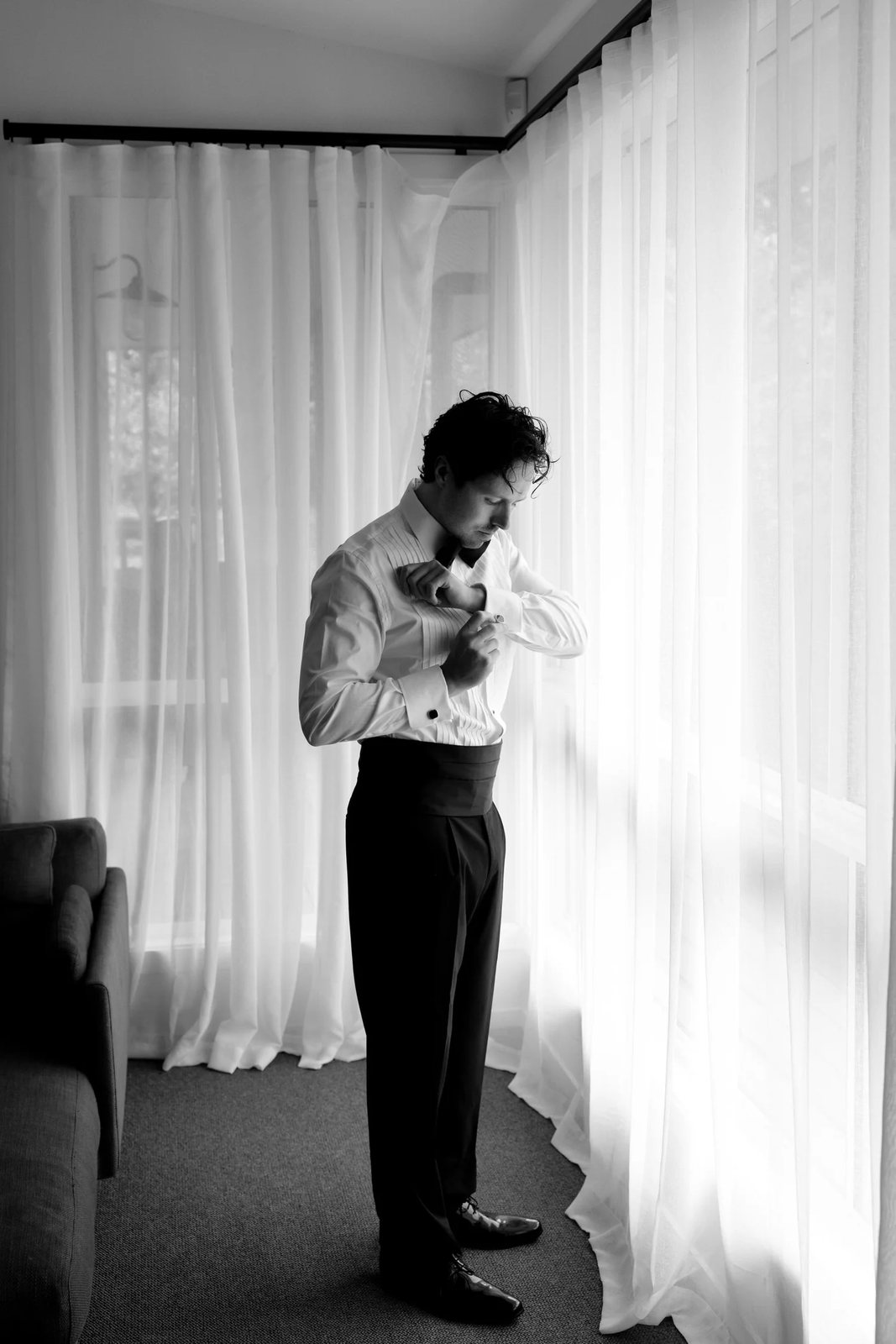 Black and white: a groom fastening his collar by a curtained window