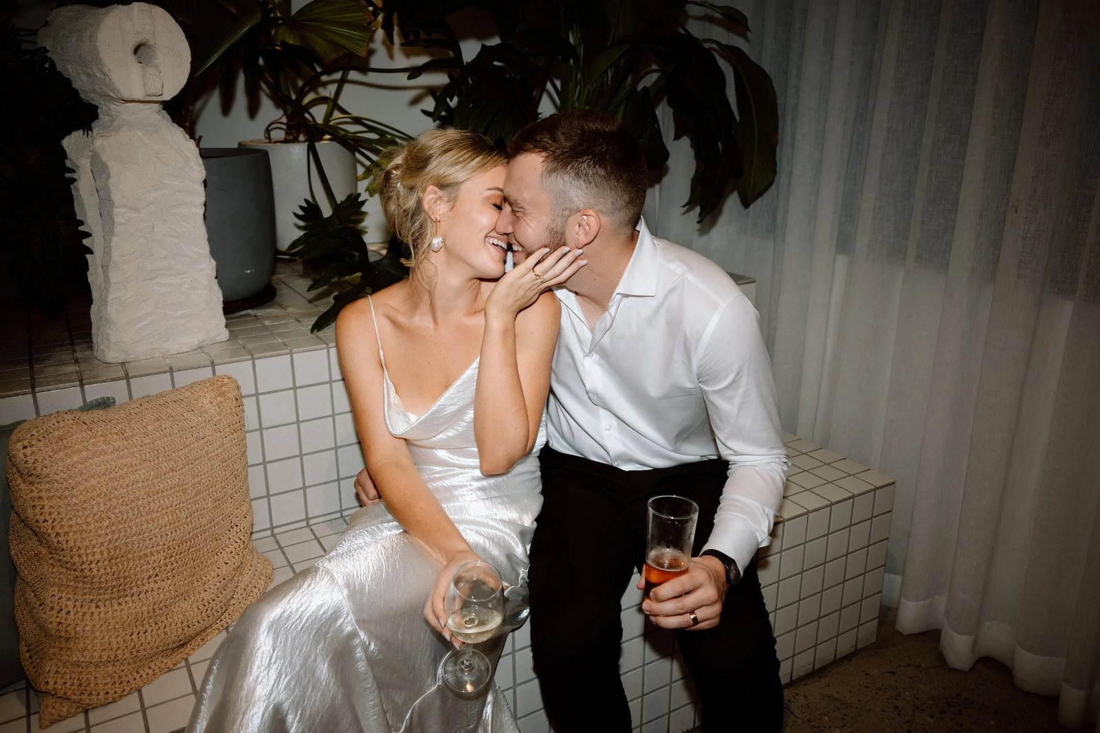 A couple sharing a kiss on a tiled bench late at night, drinks in hand