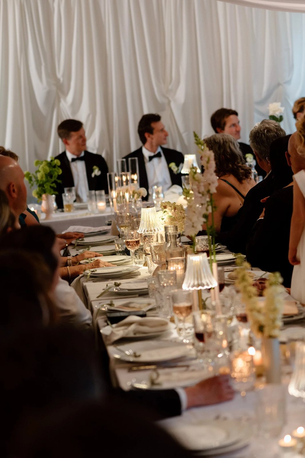 A long candlelit reception table under white drapes, guests turned towards a speech