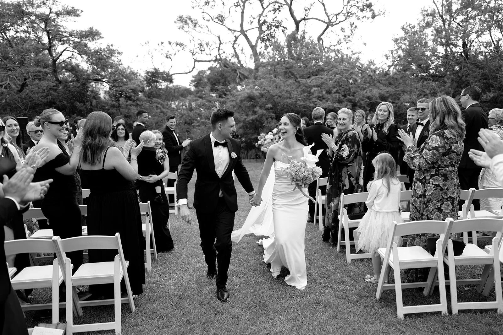 Black and white: a couple walking back up the aisle between rows of white chairs as guests clap