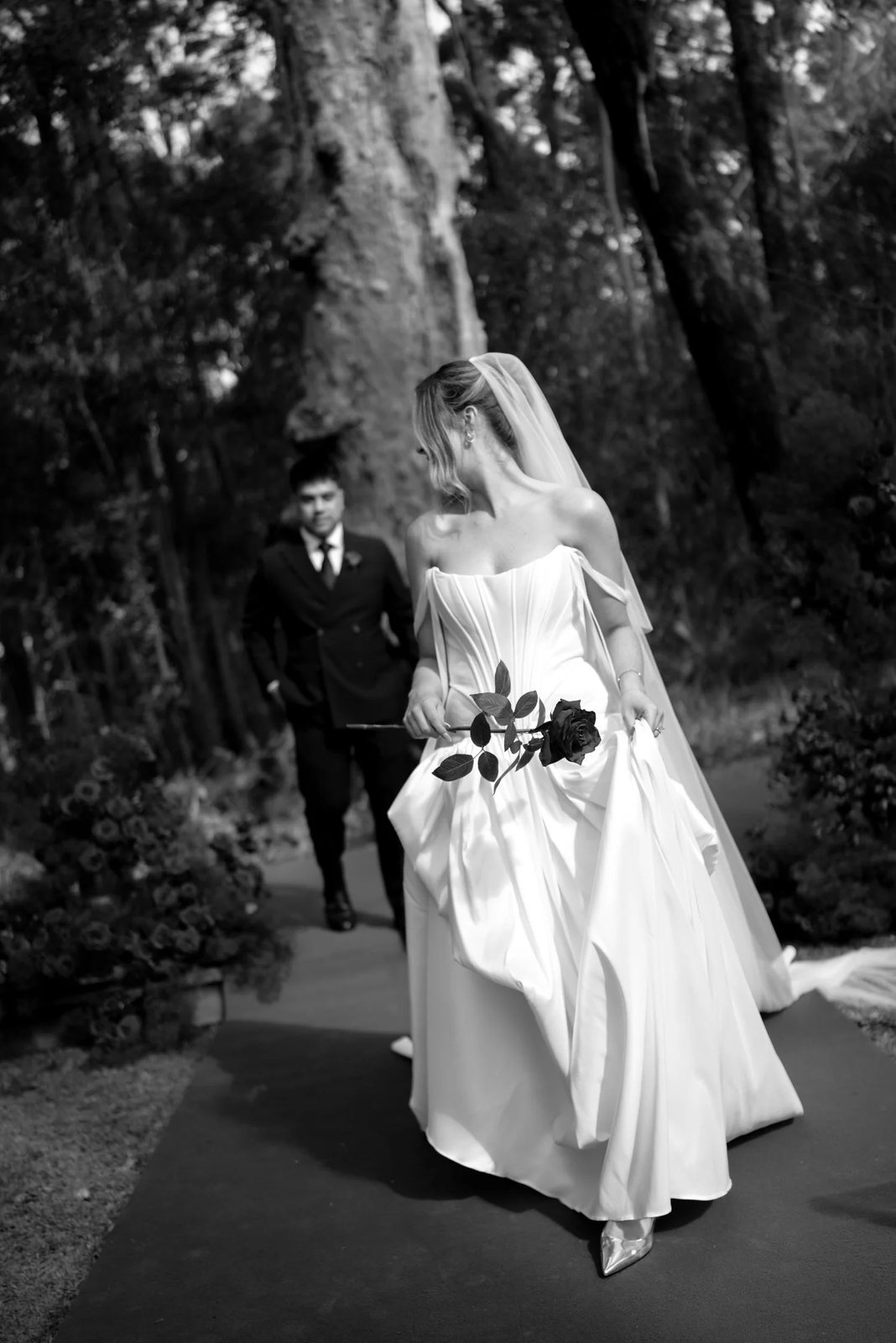 Black and white: a bride lifting her gown as she walks among tall trees, the groom behind